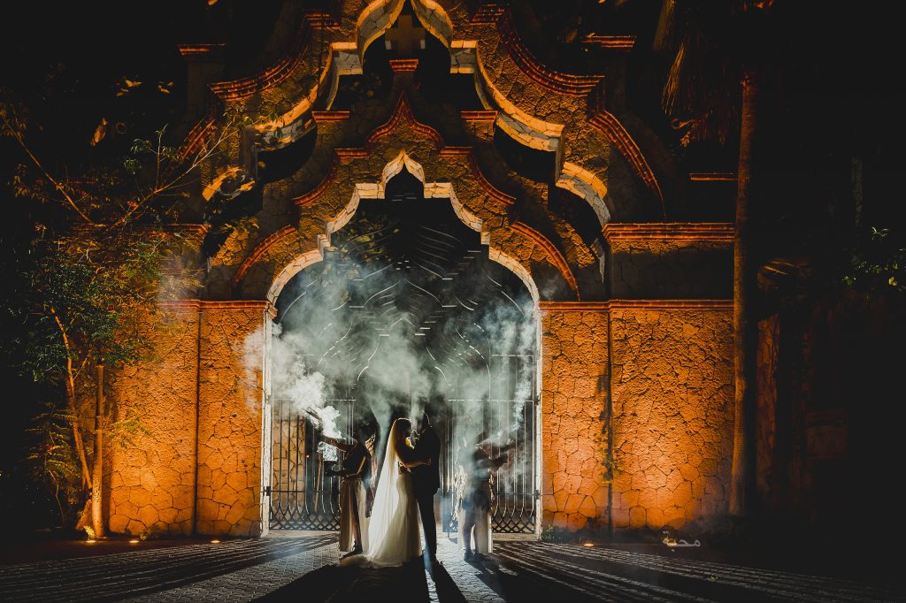 Novios abrazados de noche frente a un arco de piedra iluminado, entre el humo del sahumerio